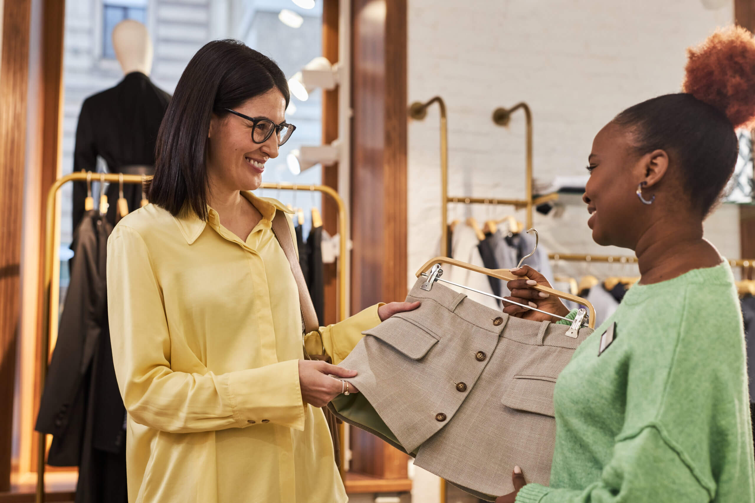 Waist up portrait of smiling woman looking at clothes in boutique with shop assistant helping